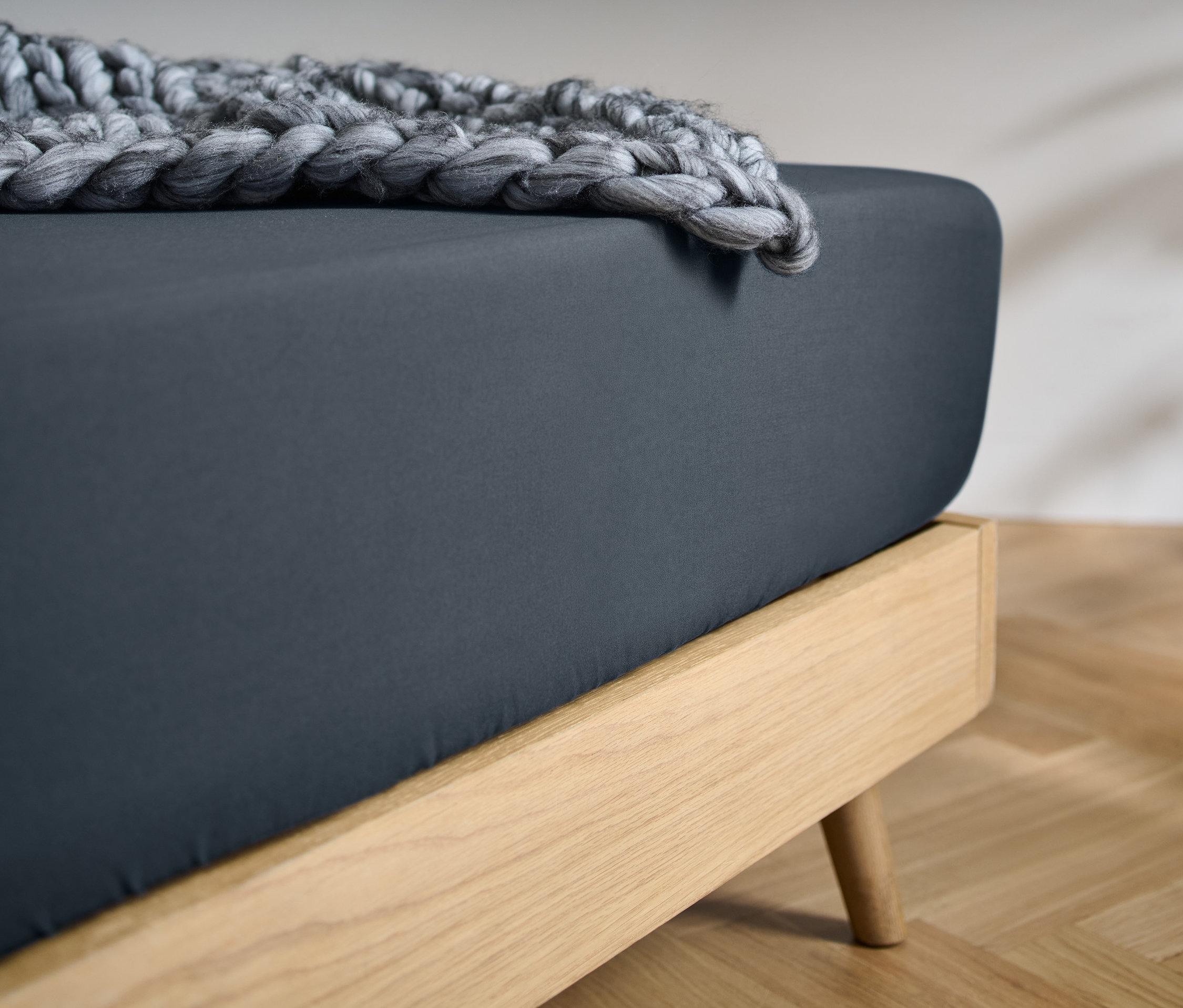 Detailní pohled na postel s tmavě modrým napínacím prostěradlem z mikrovlákna standardní velikosti a pletenou šedou dekou.