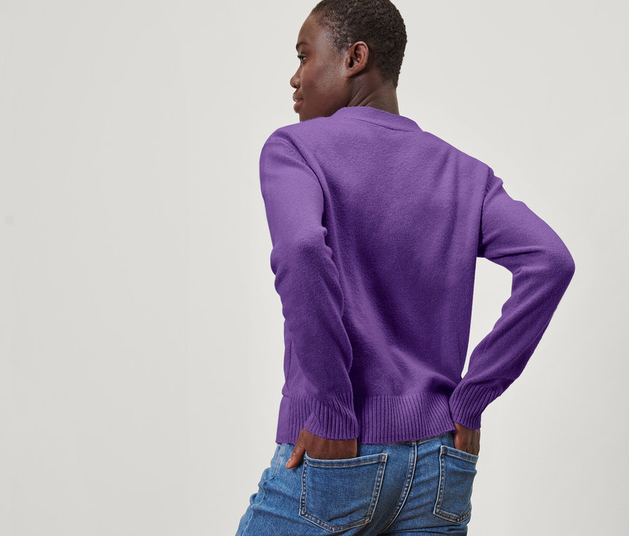 Žena pózuje zády a má ruce v kapsách džínů. Oblečená do fialového pleteného svetru s vlnou.