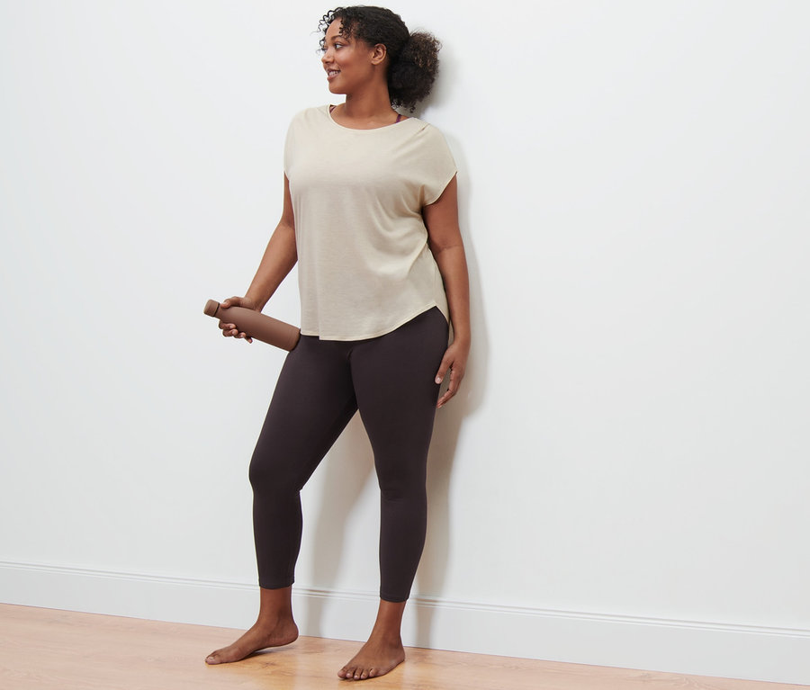 Žena drží hnědou láhev na vodu, má na sobě béžové sportovní triko a sportovní legíny.