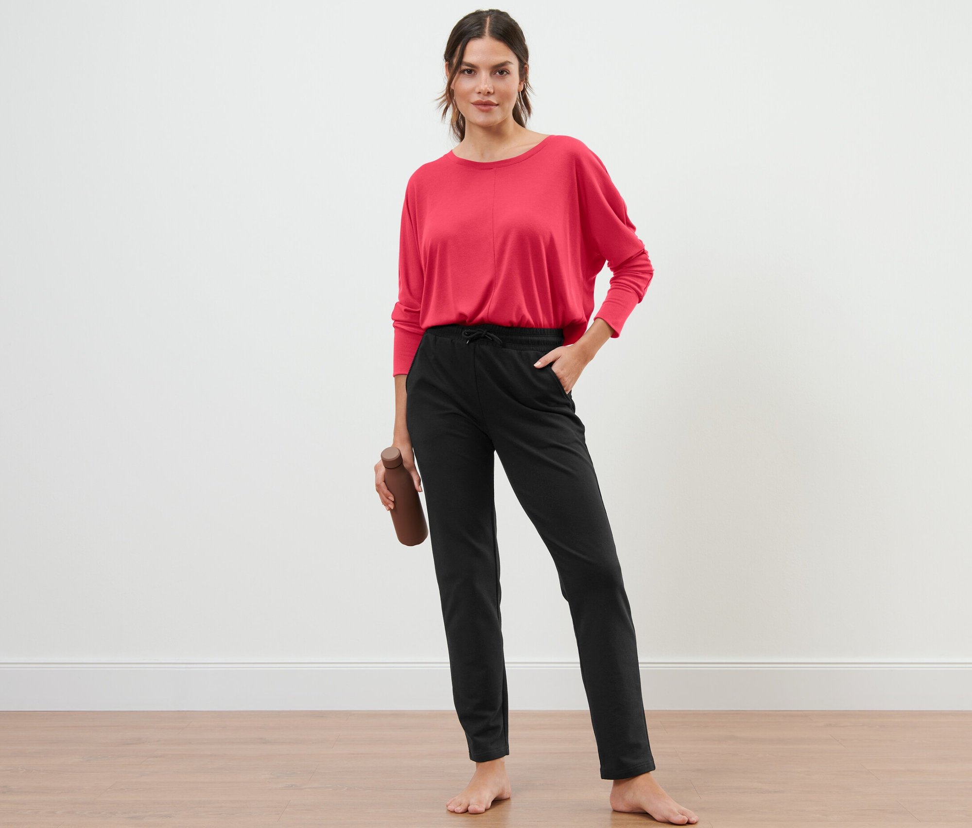 Žena stojí a drží láhev na vodu, na sobě má černé teplákové kalhoty.