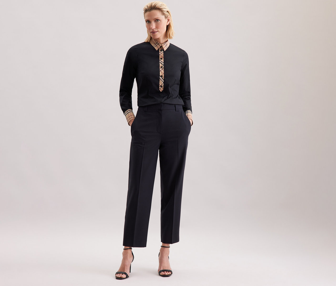 Žena pózuje v černé popelínové halence Seidensticker s kostkovaným detailem a tmavých kalhotách.