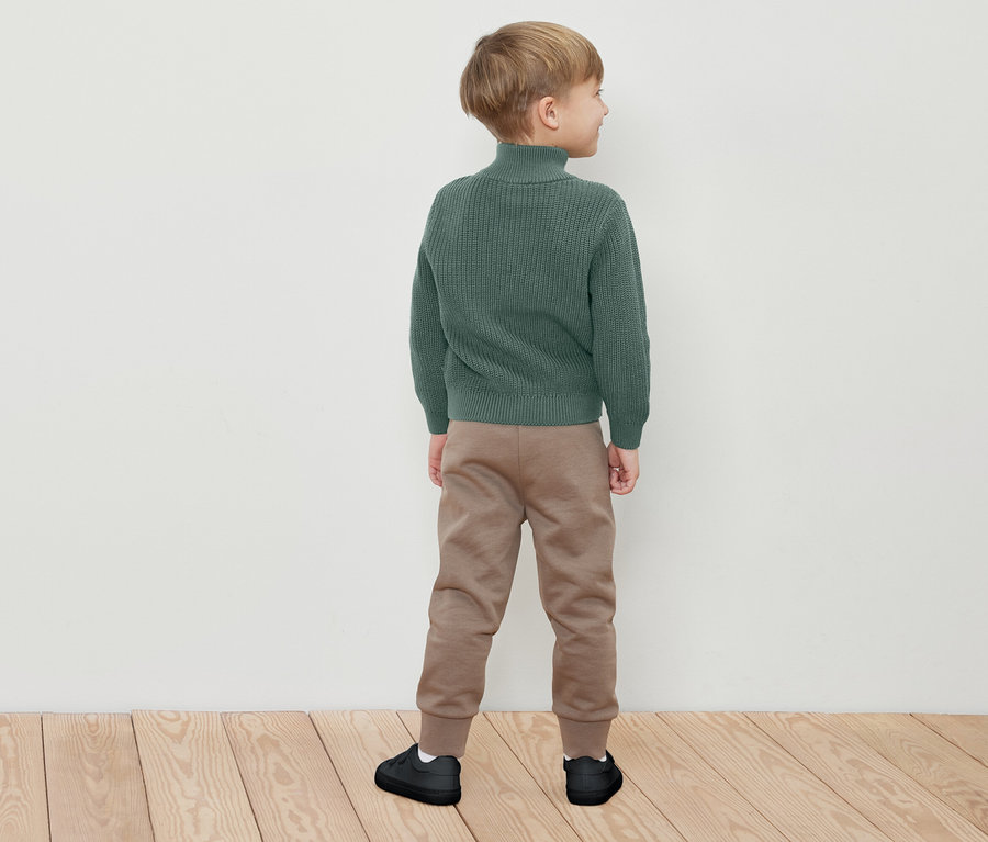 Chlapec stojí zády, má na sobě zelený rolák, hnědé tepláky a černé tenisky.