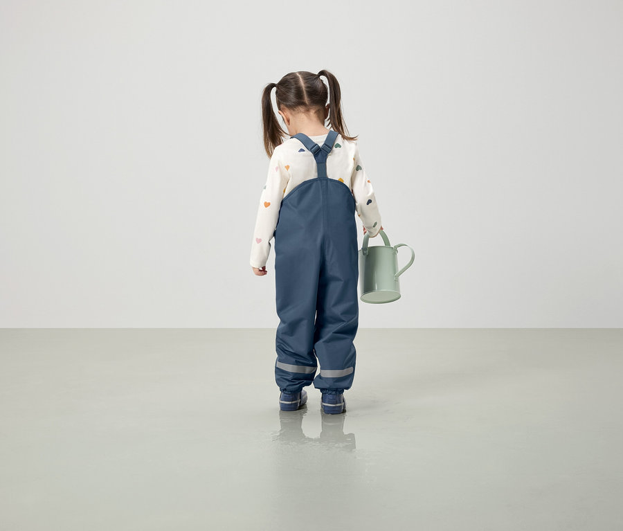 Dívka v modrých kalhotách s laclem drží zelenou konev.