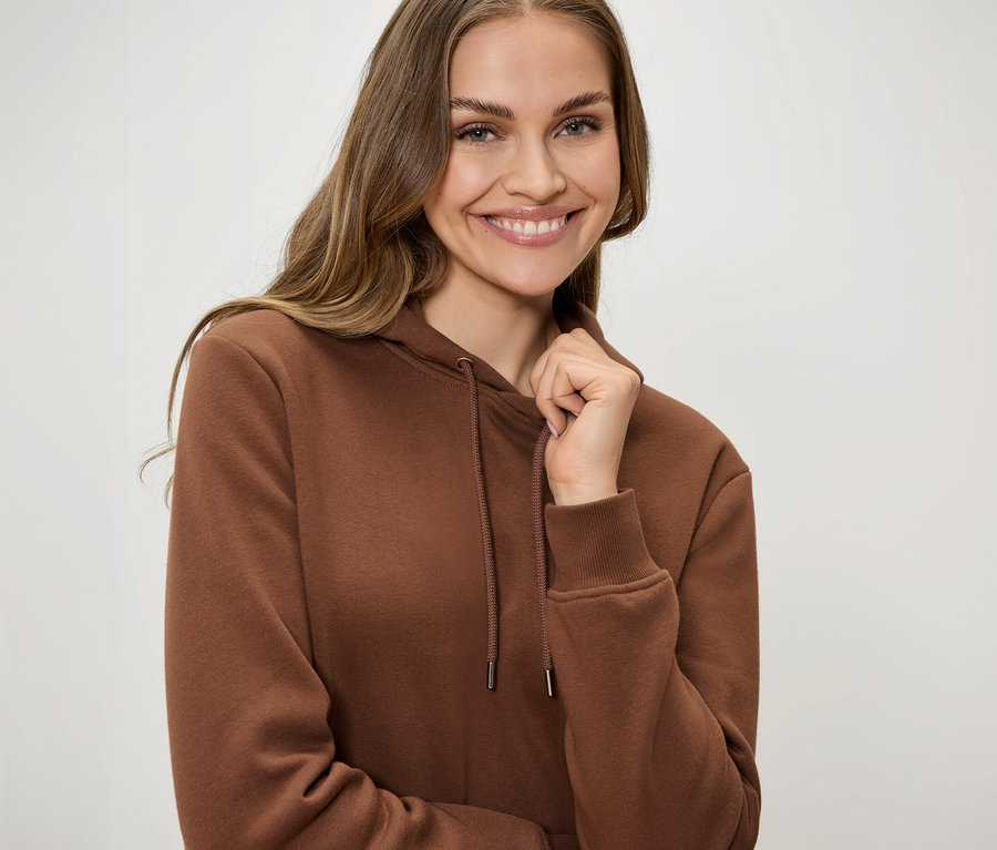 Usmívající se žena v hnědé mikině s kapucí se šňůrkami drží límec jednou rukou.
