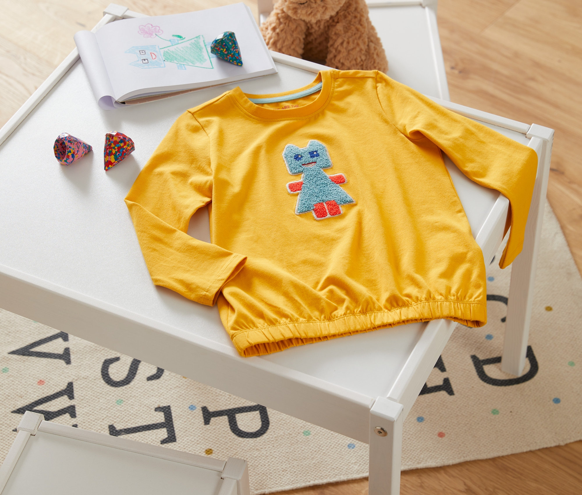 Žluté triko s dlouhým rukávem leží na bílém stole vedle otevřeného zápisníku a pastelek. Na židli sedí plyšový pes.