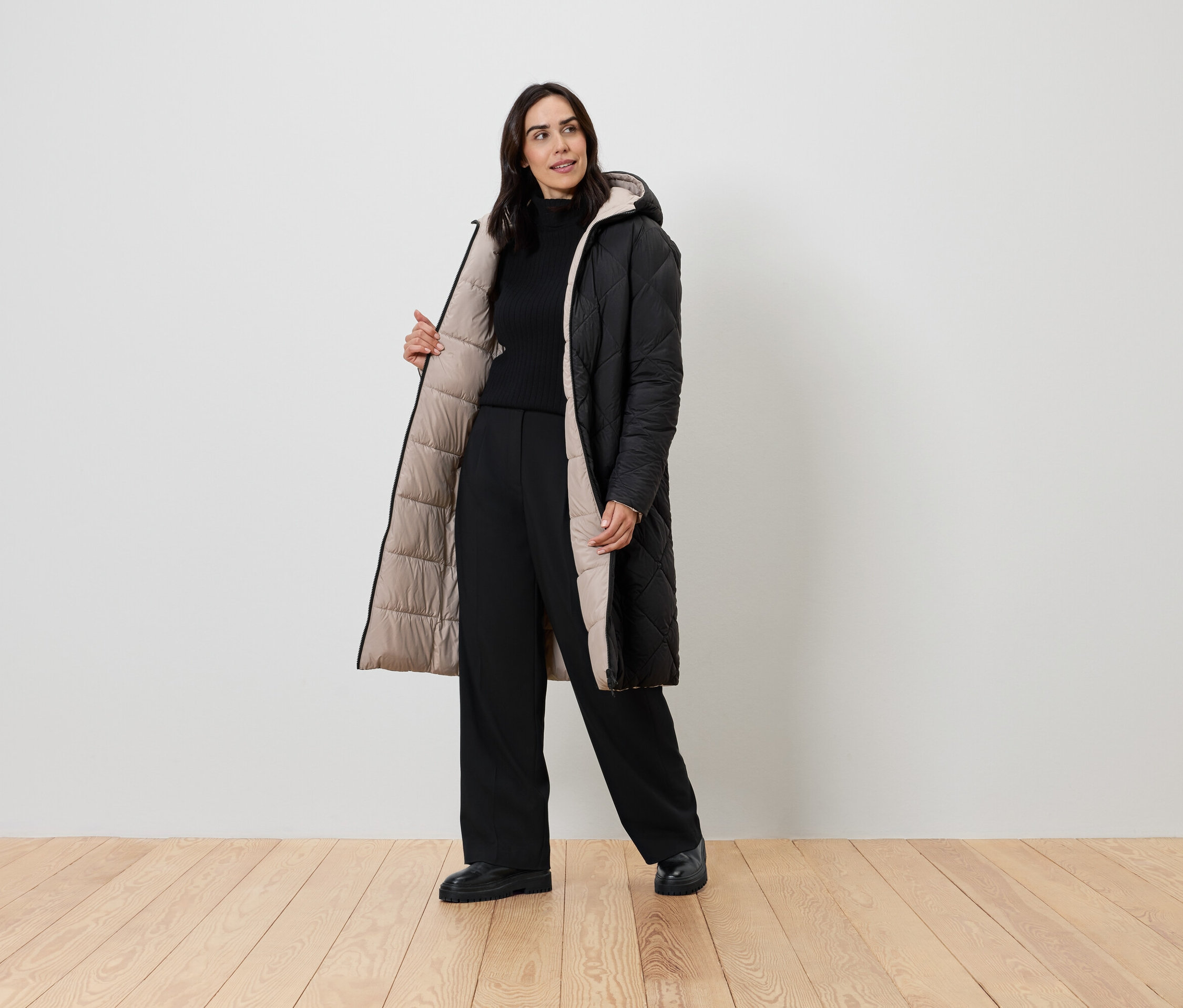 Žena pózuje v černém kabátě s béžovou podšívkou, černém roláku a černých kalhotách.