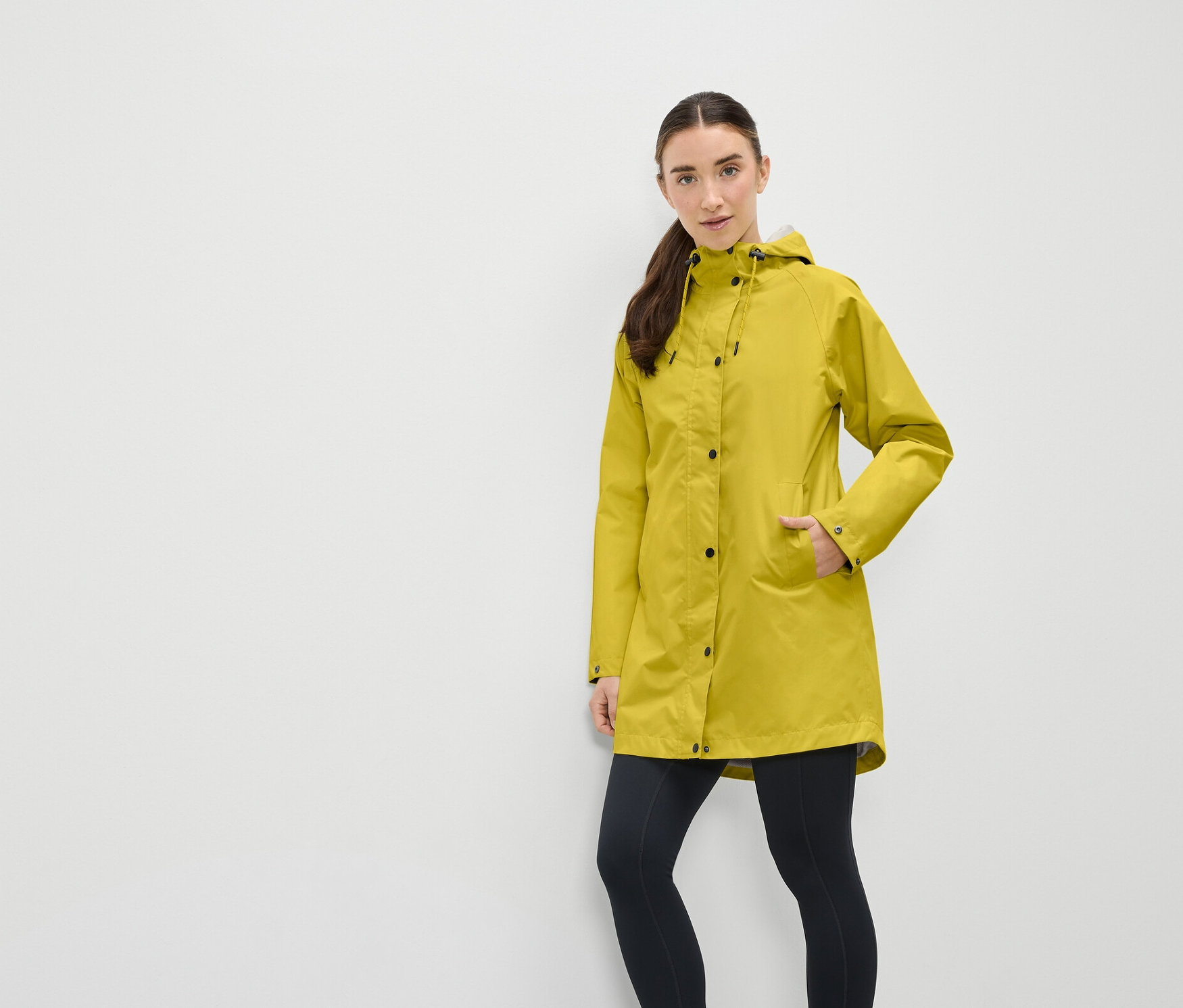 Žena v žlutém kabátu s kapucí a černých legínách stojí před bílou zdí.
