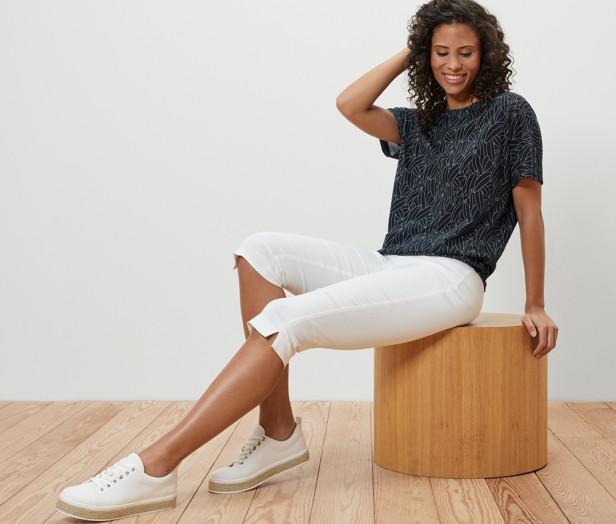 Žena sedí na stoličce a má na sobě tmavě modré halenkové tričko se spadlými rameny, bílé capri kalhoty a tenisky.