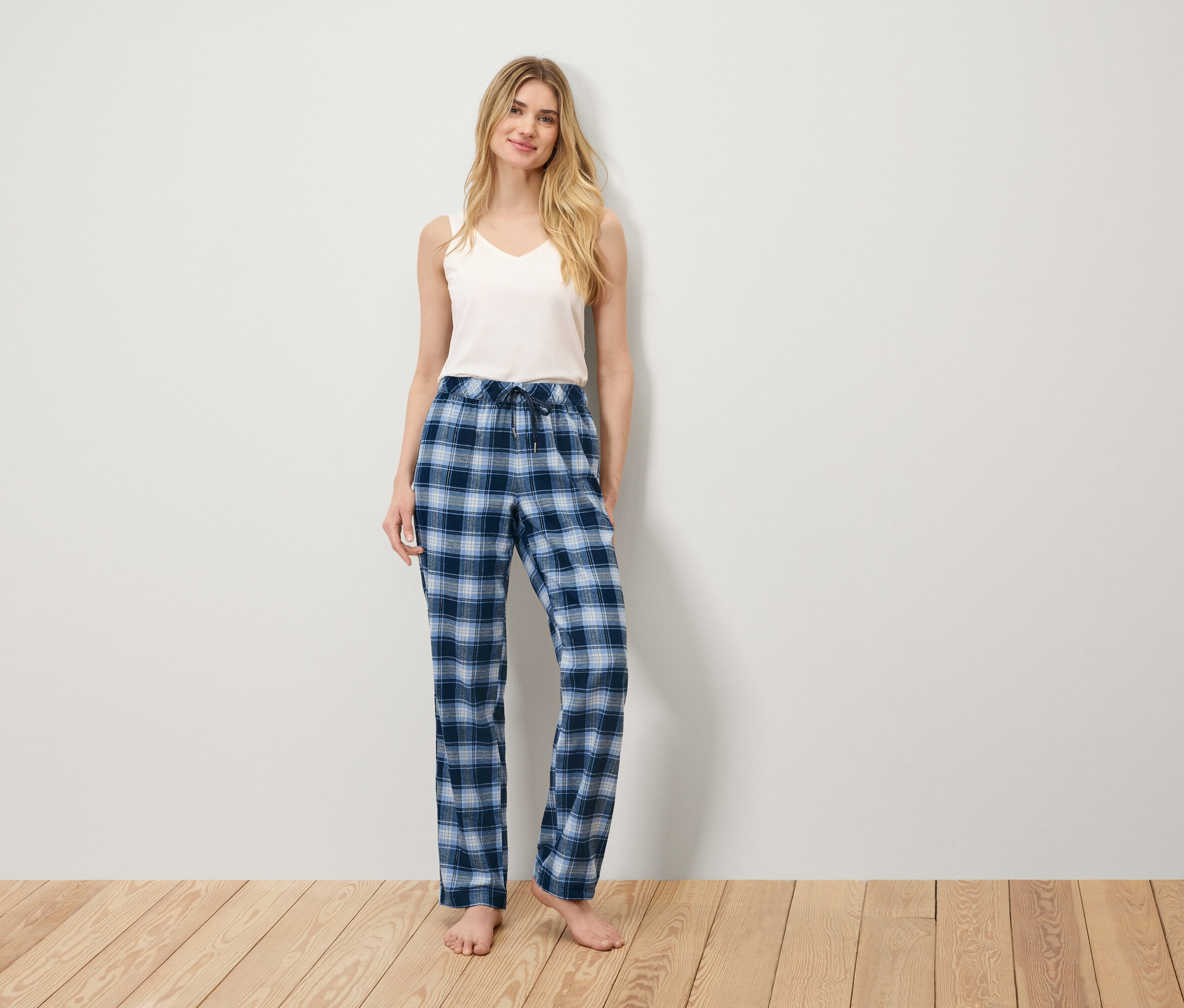 Žena stojí na dřevěné podlaze a má na sobě bílé tílko a modré kostkované pyžamové kalhoty.
