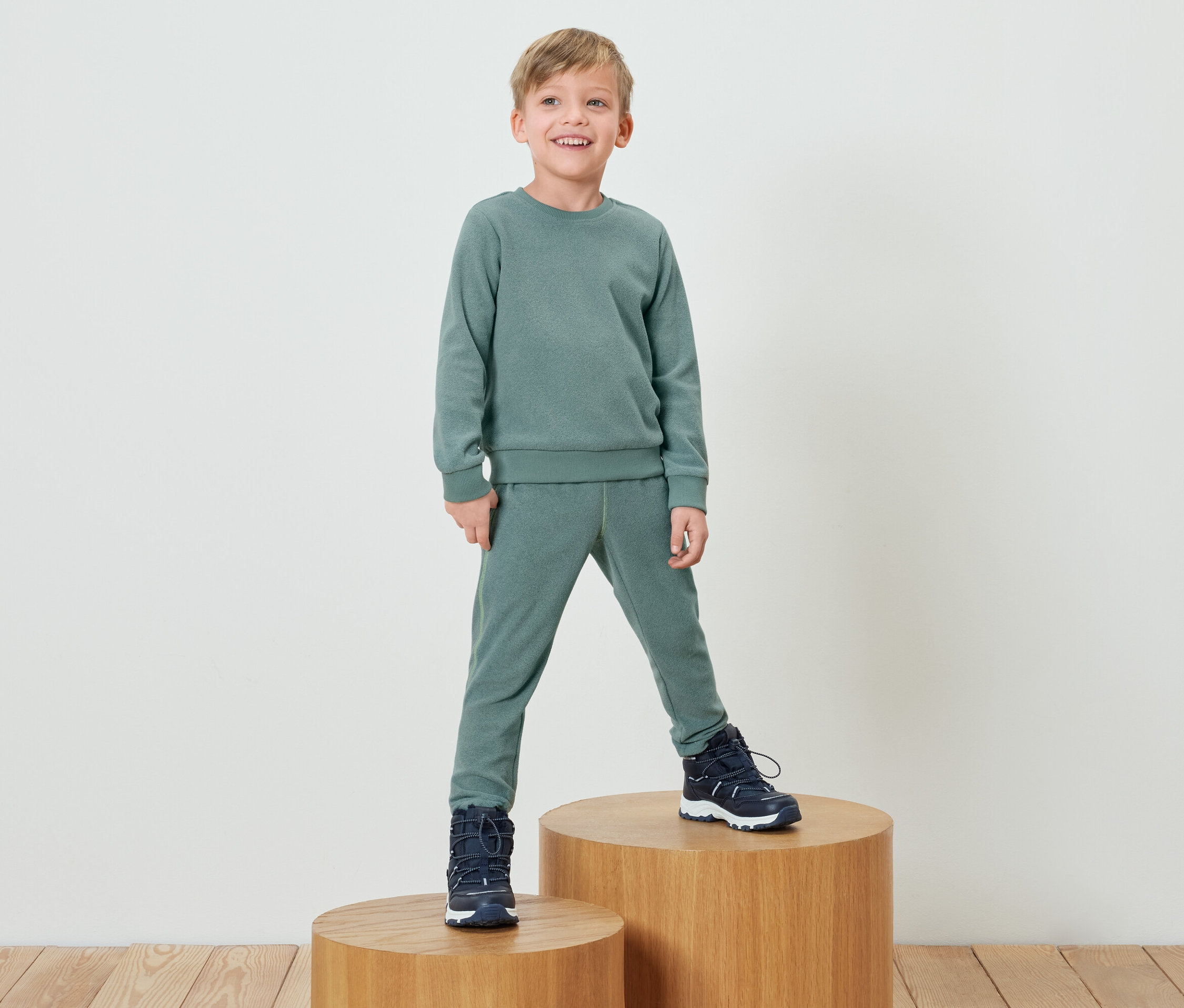 Chlapec v zelené soupravě stojí na dřevěném podstavci.