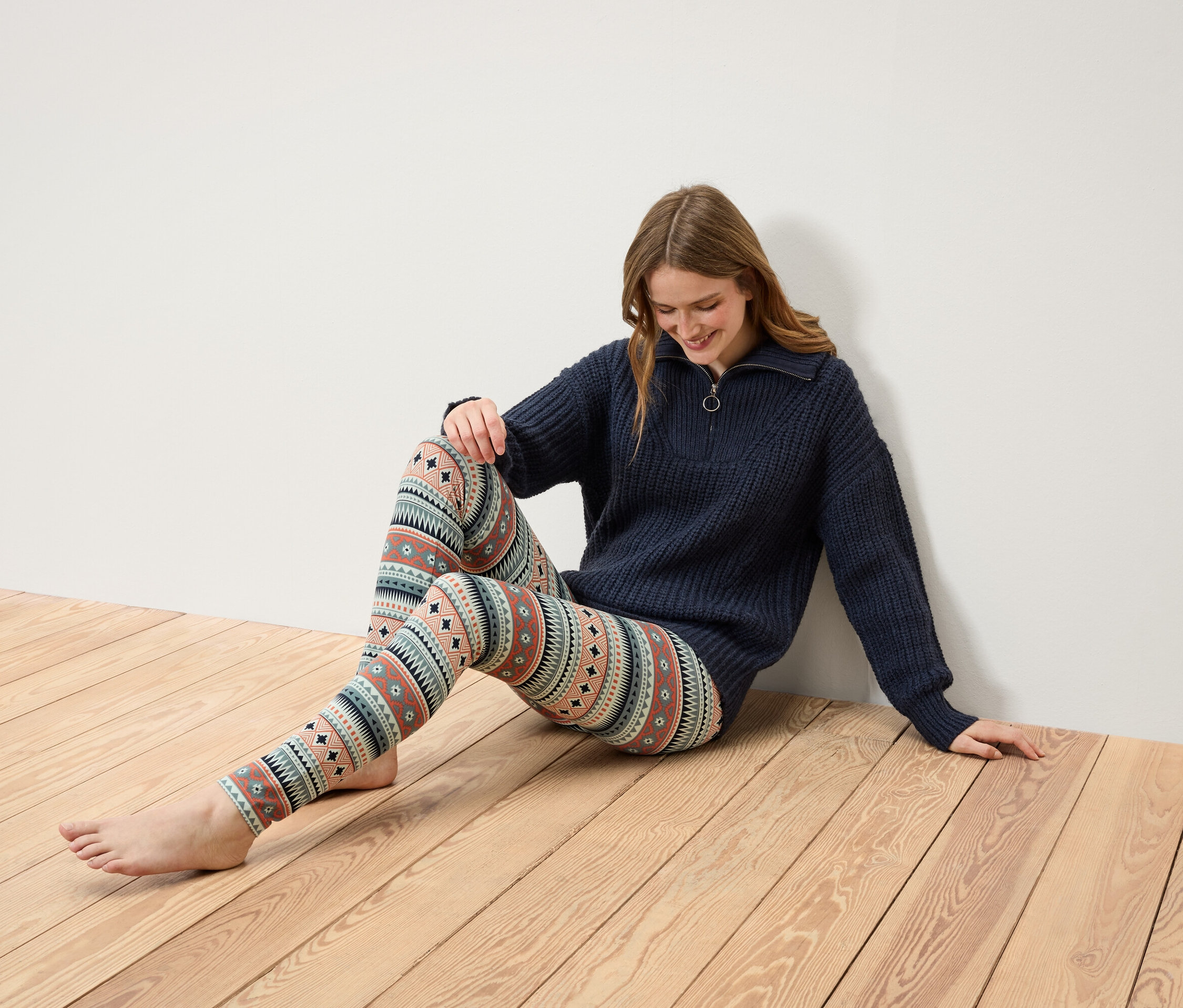 Žena sedí na dřevěné podlaze, má na sobě tmavě modrý svetr na zip a vzorované legíny.