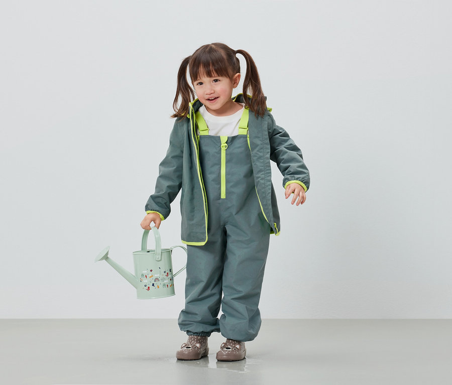 Dívka v šedých nepromokavých kalhotách a bundě drží světle zelenou konvičku.