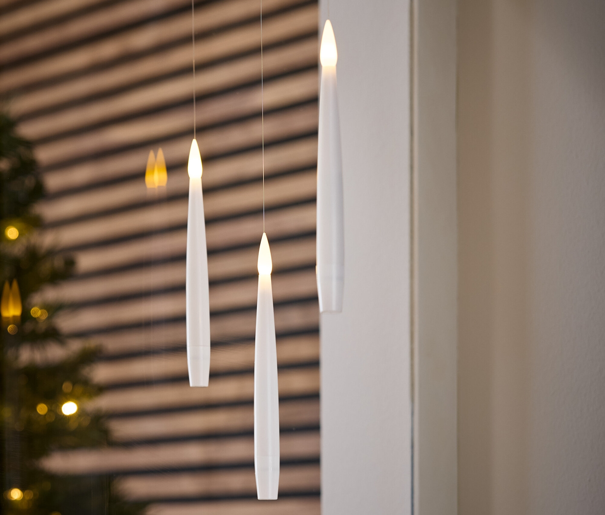 Tři bílé, elektronické svíčky visí na provázcích. V pozadí je vánoční stromek a dřevěná zeď.