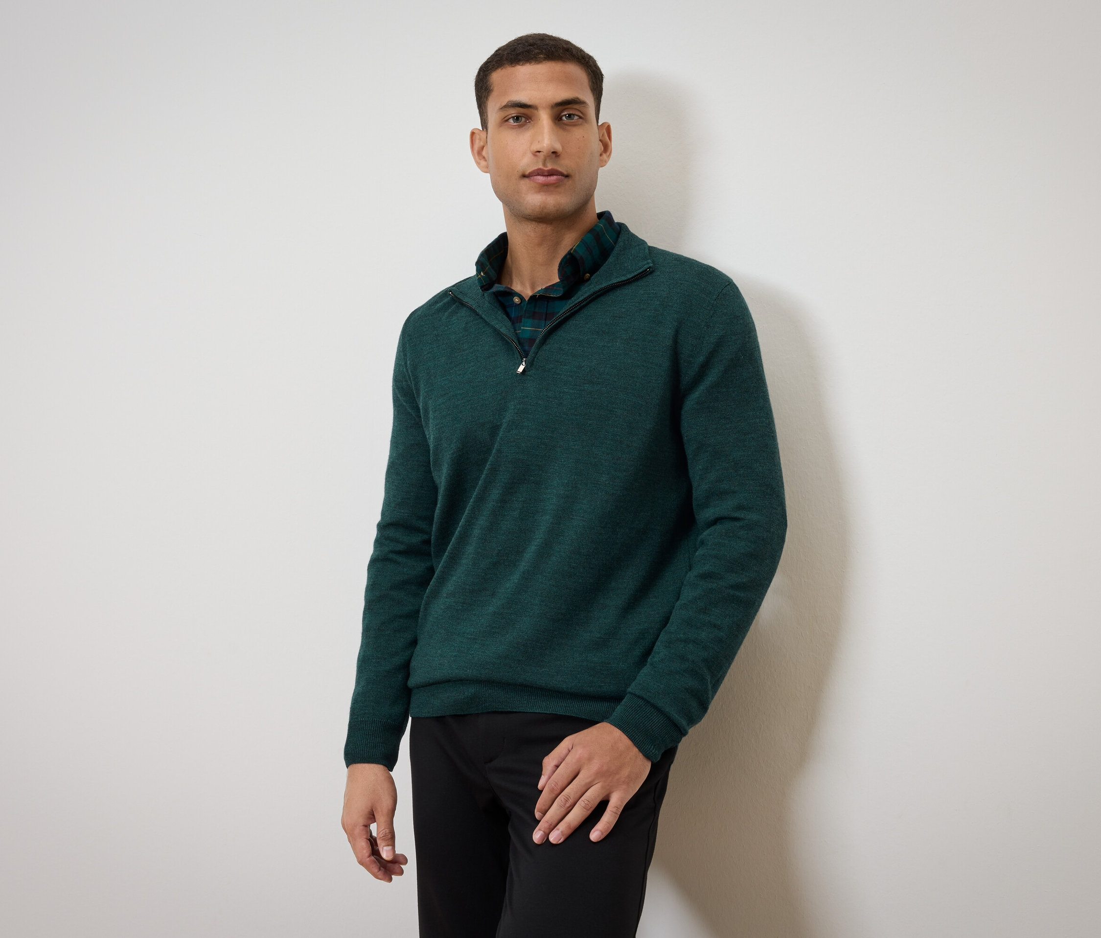 Muž pózuje v zeleném svetru a černých kalhotách.