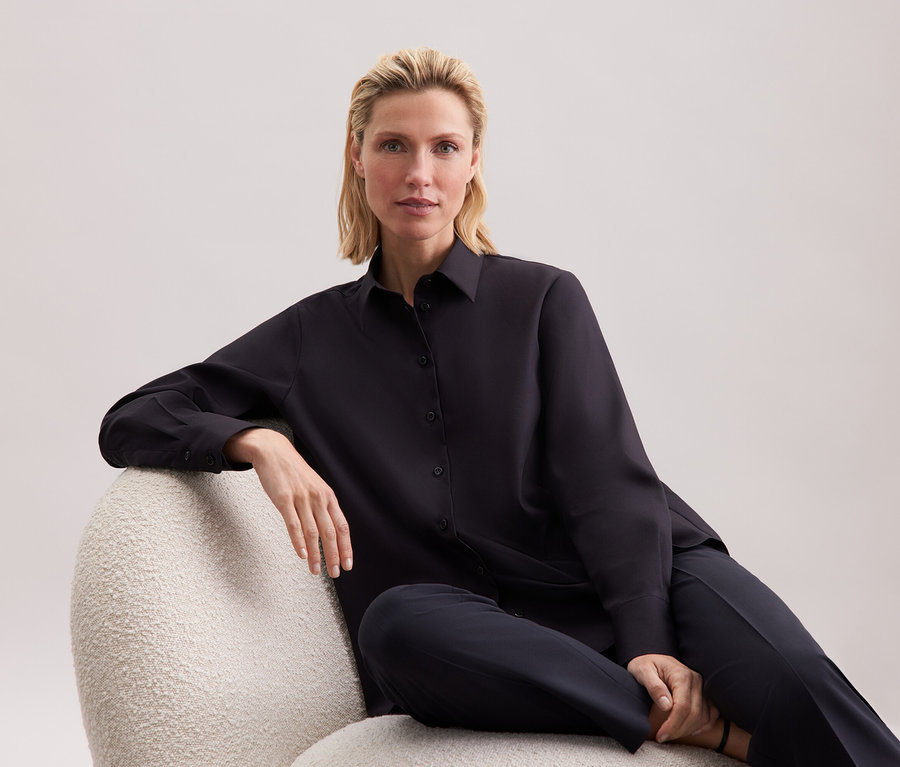 Blondýna sedí v křesle v černé košili a kalhotách.