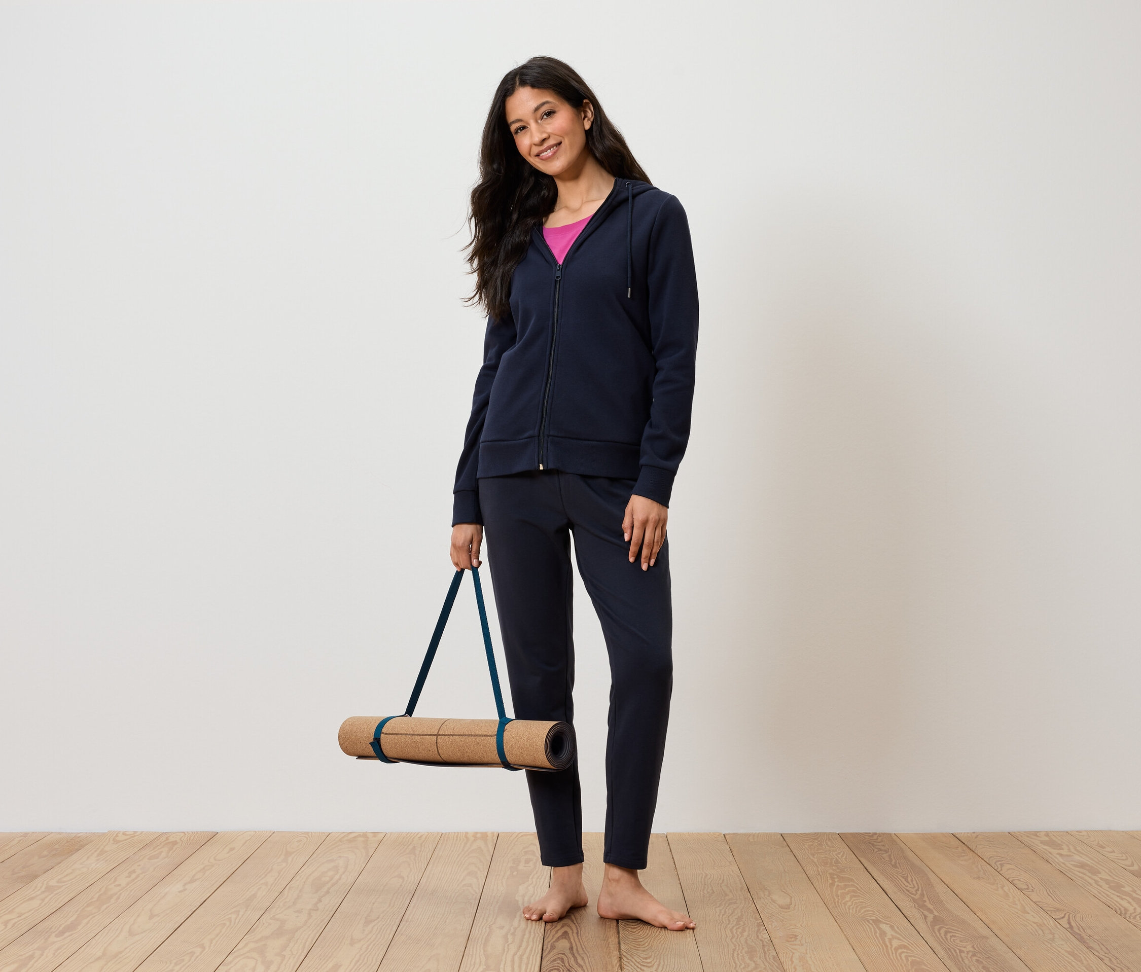 Žena drží podložku na cvičení jógy, oblečená v tmavě modré soupravě a růžovém tílku.