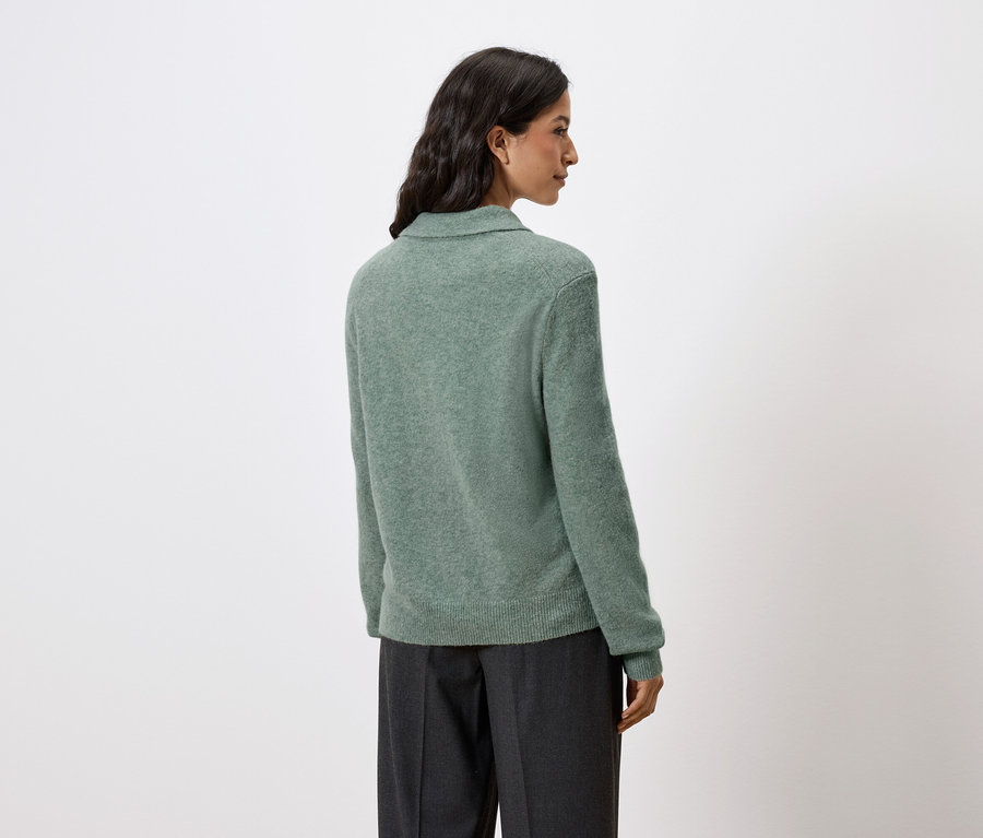 Zezadu je vidět žena s tmavými vlasy, která má na sobě zelený svetřík a tmavé kalhoty.