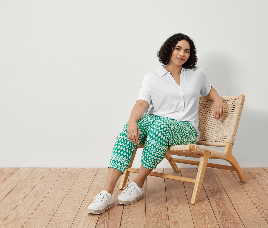 Žena sedí v křesle a má na sobě bílou halenku s knoflíkovou légou a 3/4 žerzejové kalhoty s potiskem.