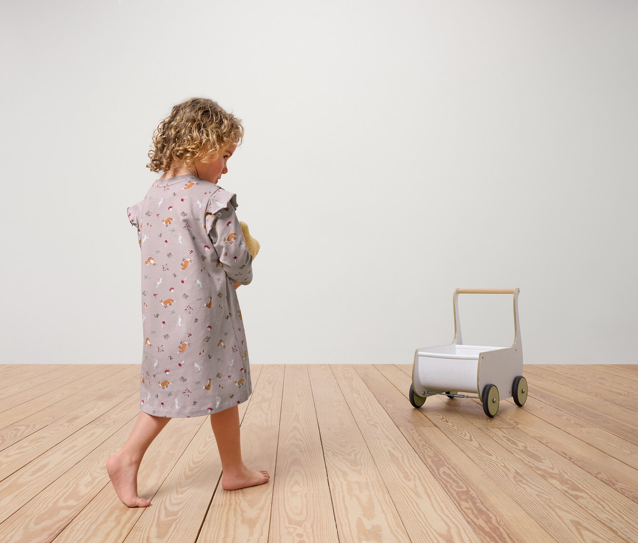 Dívka zezadu ve 2dílné dětské noční košili drží plyšovou hračku. Na dřevěné podlaze stojí vozík na kolečkách.