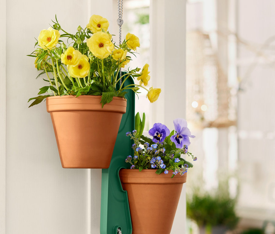Dva terakotové květináče se žlutými a modrými květy visí na zeleném držáku na bílých dveřích.