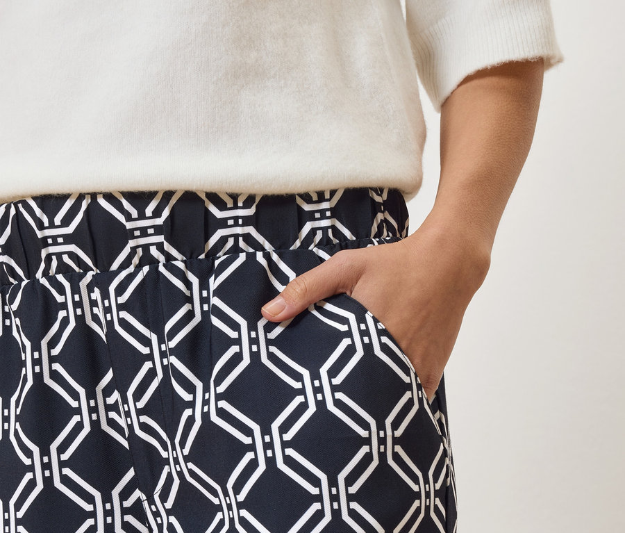 Detail osoby v tmavě modrých kalhotách s bílým geometrickým vzorem a bílém svetru, s rukou v kapse.