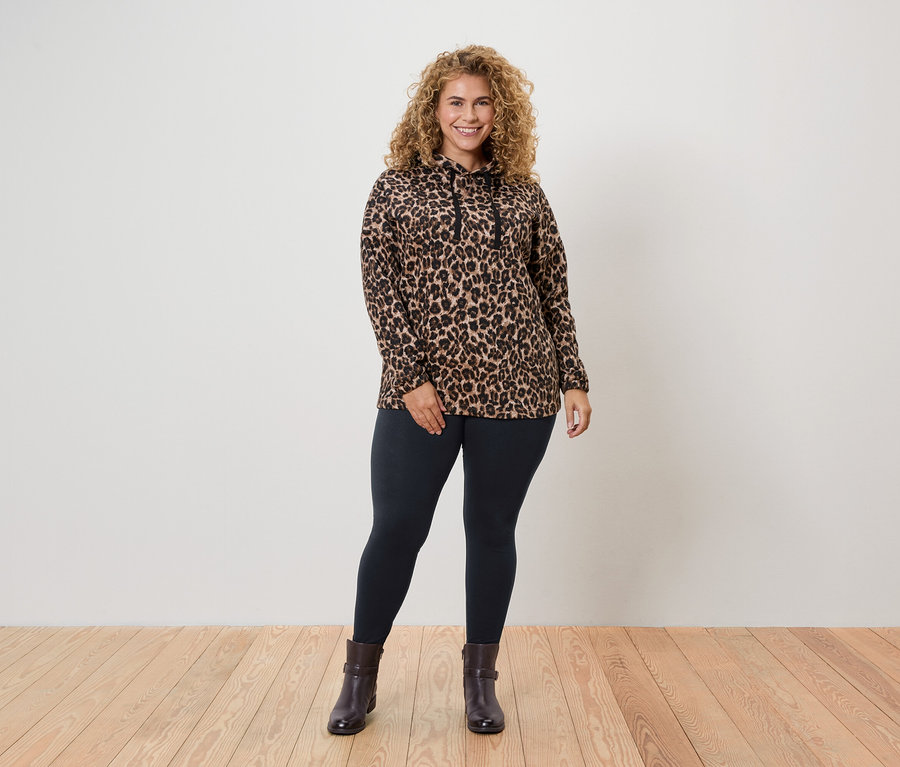 Žena s kudrnatými vlasy stojí a má na sobě top s leopardím vzorem, legíny a kotníkové boty.
