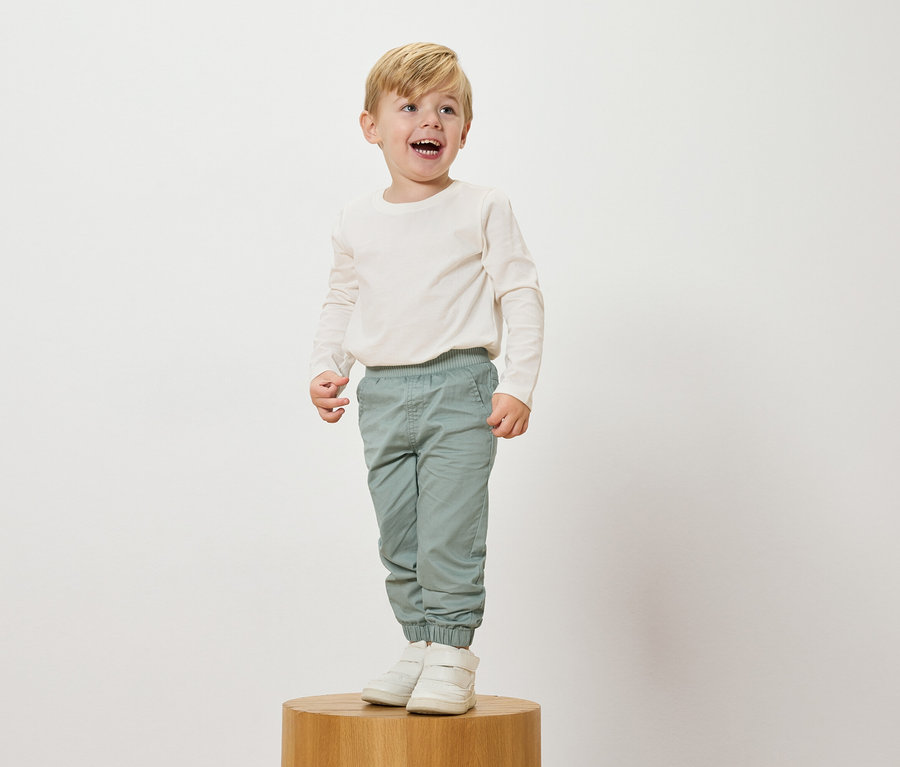 Usmívající se blonďatý chlapec stojí na dřevěném podstavci, má na sobě krémový top s dlouhým rukávem, světle modré kalhoty a bílé tenisky.