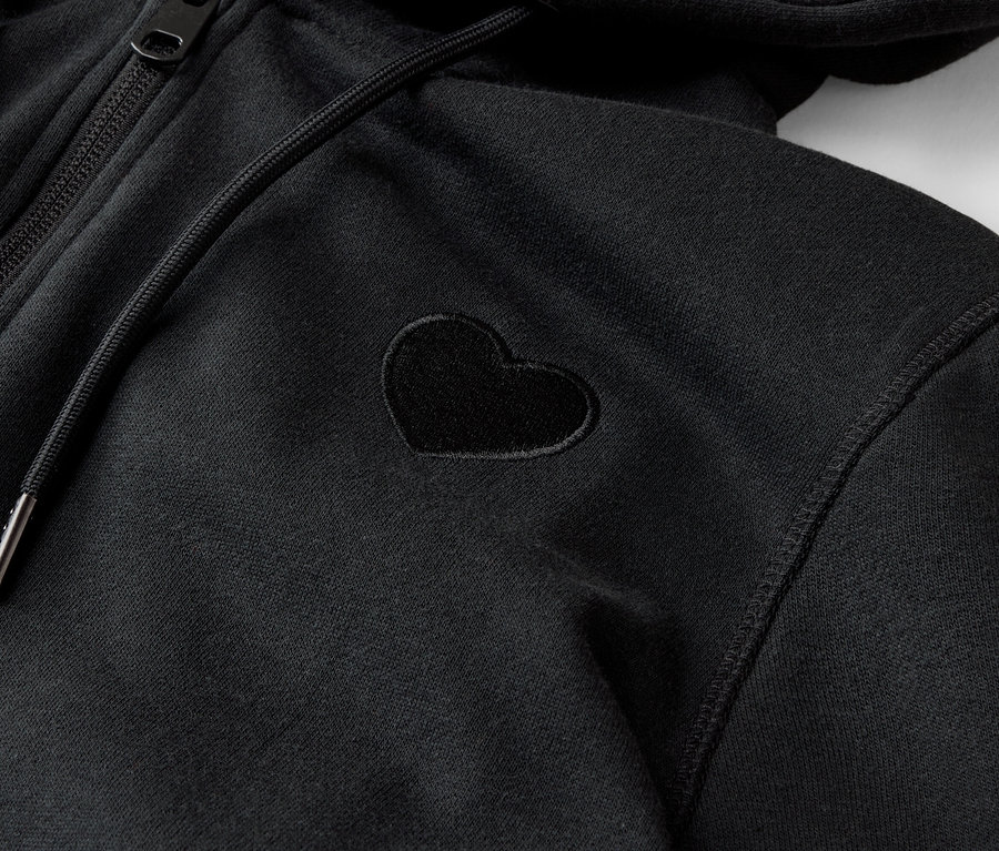 Detailní záběr na černou mikinu s kapucí na zip s malou aplikací ve tvaru srdce.