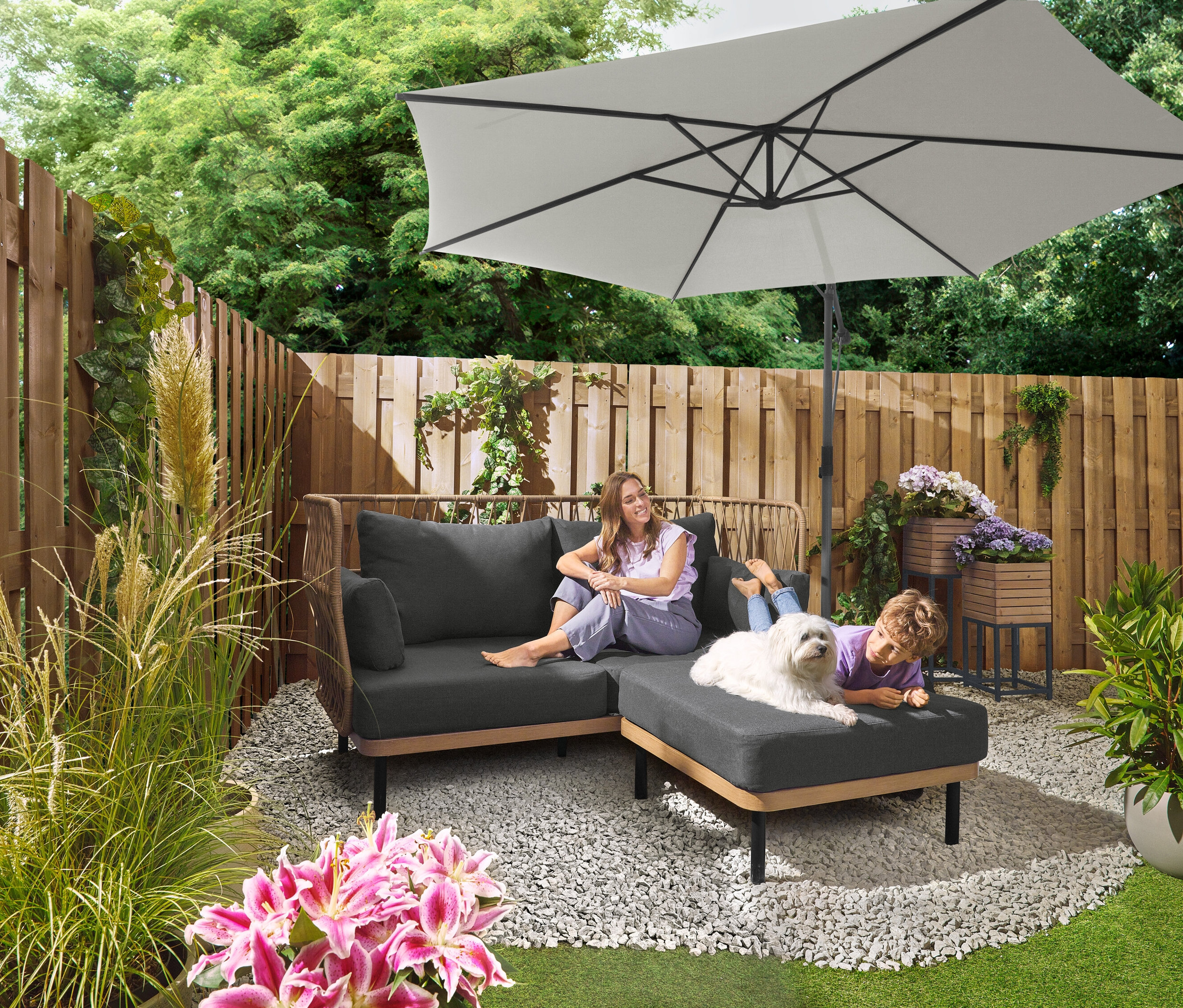 Žena sedí na šedé pohovce pod slunečníkem Leco Ampelschirm Ø 300 cm se stojanem a krytem. Dítě leží se psem na otomane. V pozadí je vidět sada desek Leco pro křížový stojan.