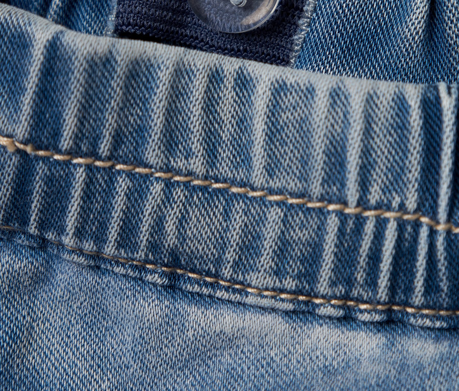 Detailní záběr na světle modré džíny s nastavitelným elastickým pasem a průhledným knoflíkem.