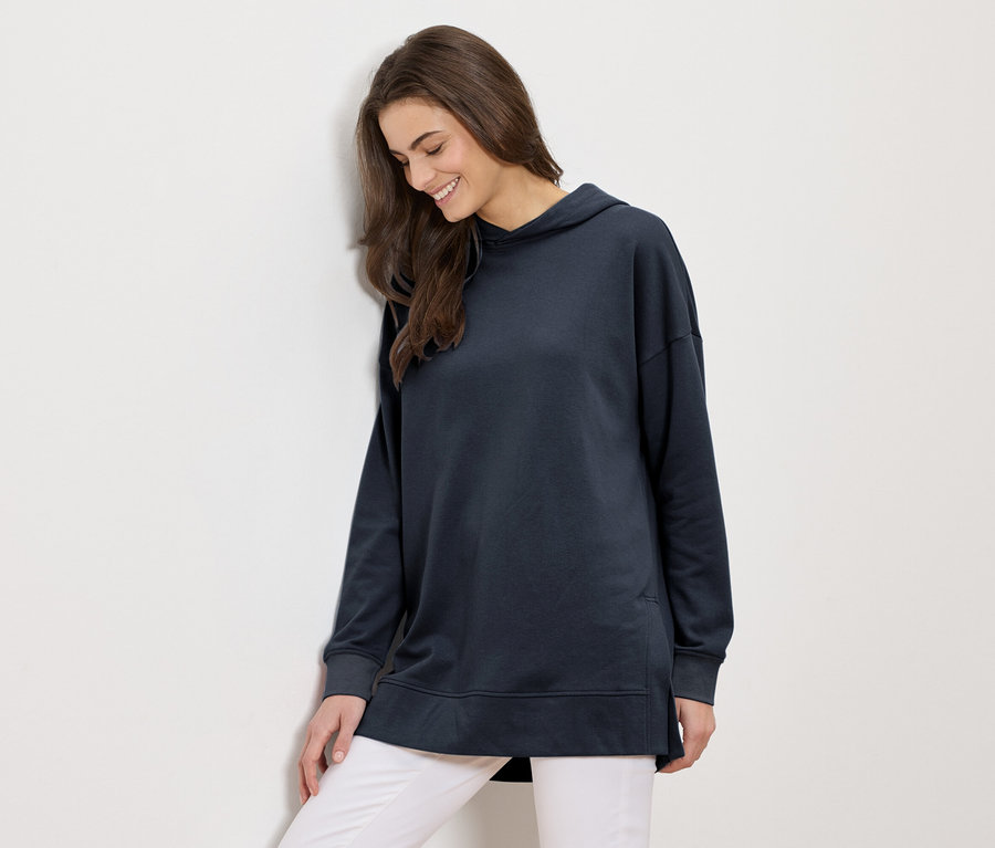 Žena s dlouhými hnědými vlasy v dlouhé tmavě modré mikině s kapucí a bílých kalhotách se usmívá a opírá se o bílou stěnu.