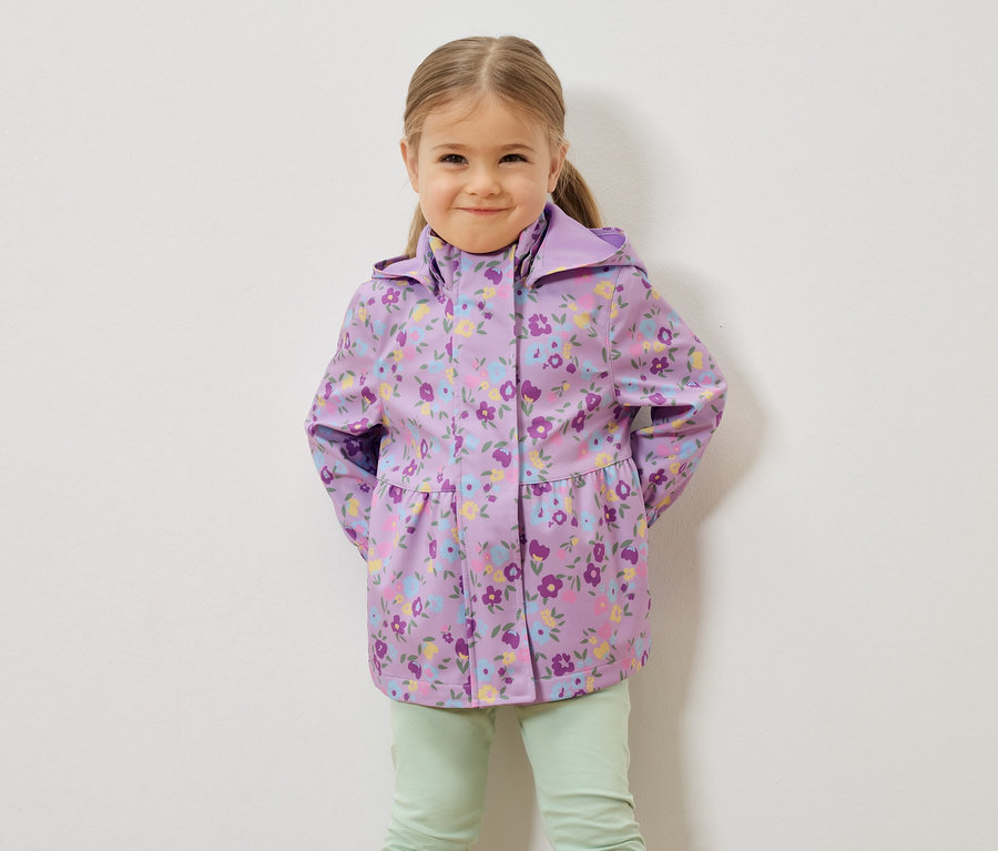 Dívka s blond vlasy má na sobě fialovou květovanou bundu do deště a světle zelené kalhoty před bílou stěnou.