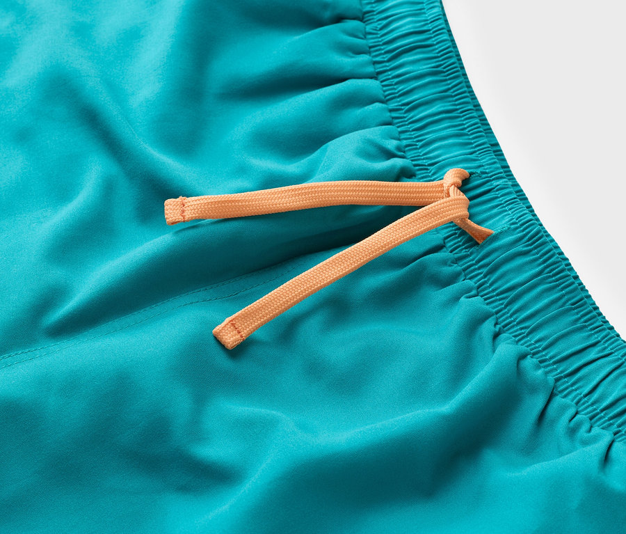 Detailní záběr na tyrkysové plavkové šortky s oranžovou šňůrkou.