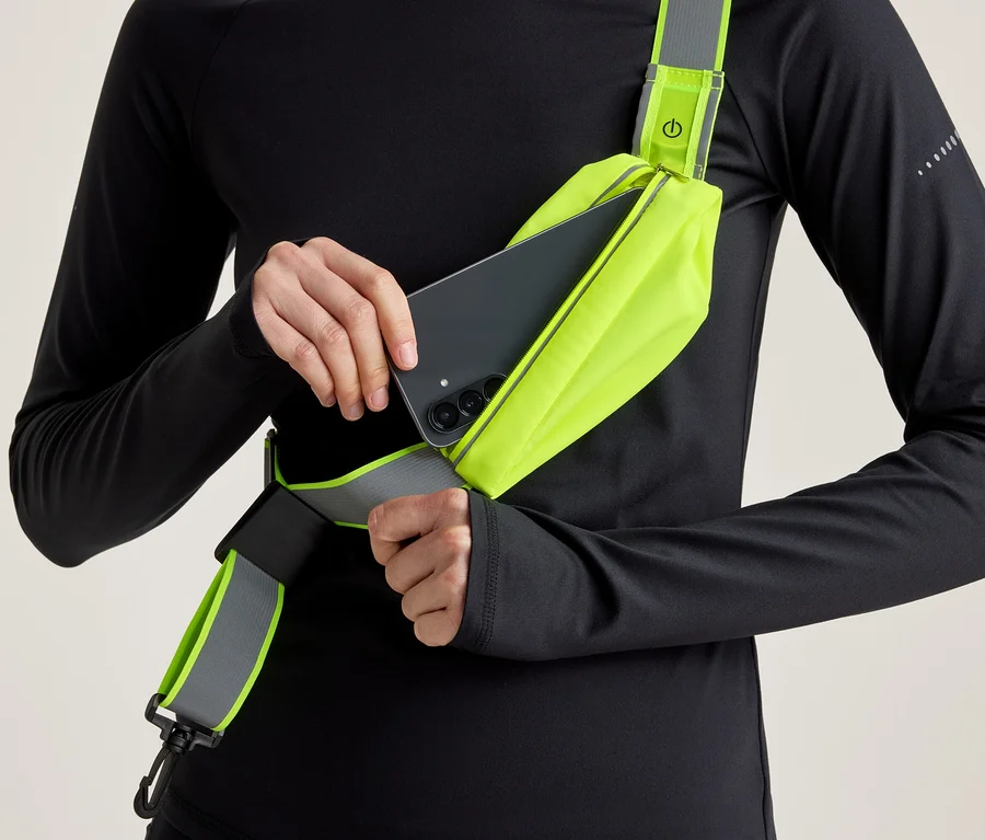 Osoba v černém tričku s dlouhým rukávem vkládá černý smartphone do neonově žluté crossbody tašky s reflexním detailem.