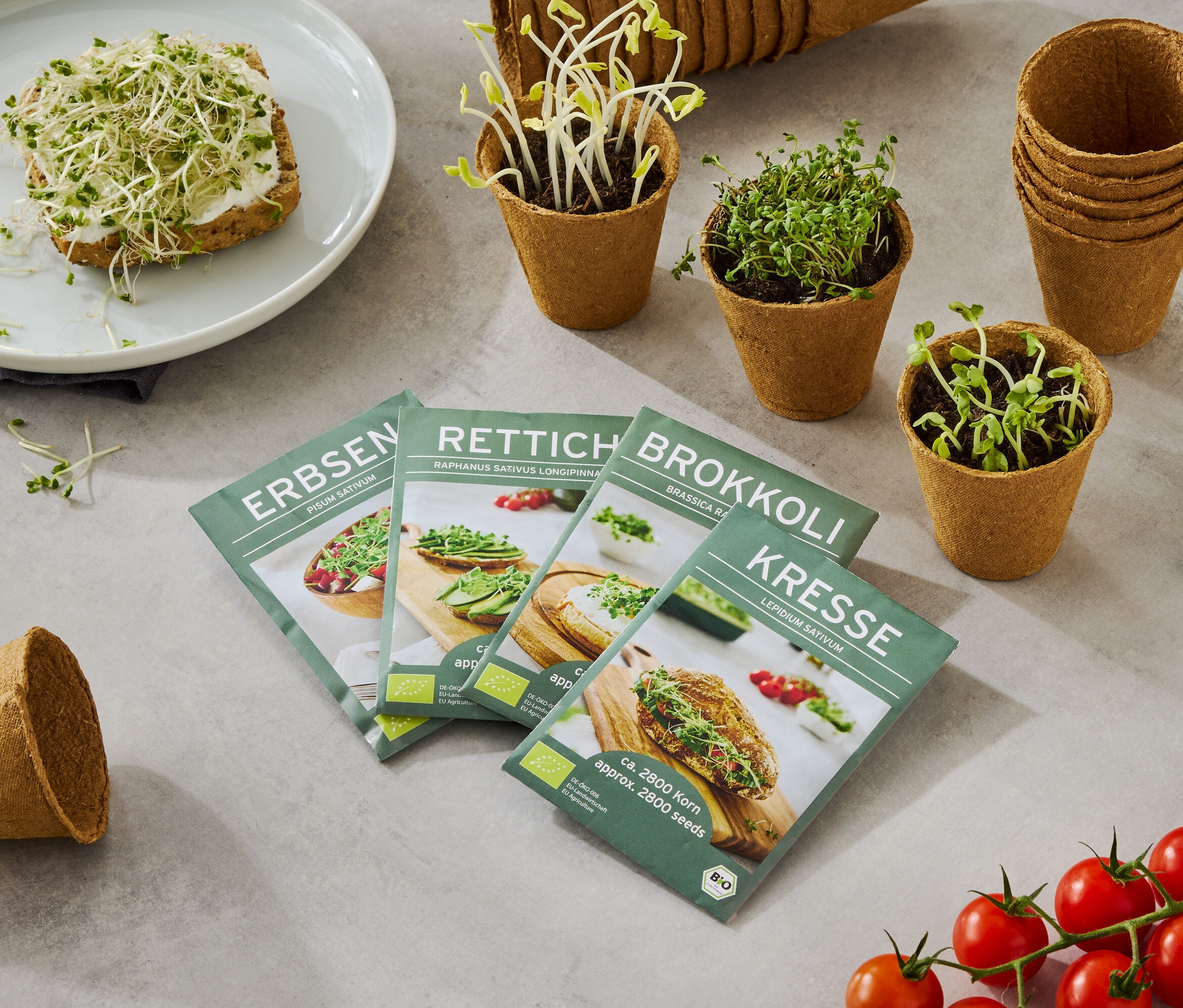 Talíř s chlebem posypaným klíčky, spolu se čtyřmi krabicemi semen (hrách, ředkvička, brokolice, řeřicha), malými květináči s rostoucími klíčky a trsem rajčat.