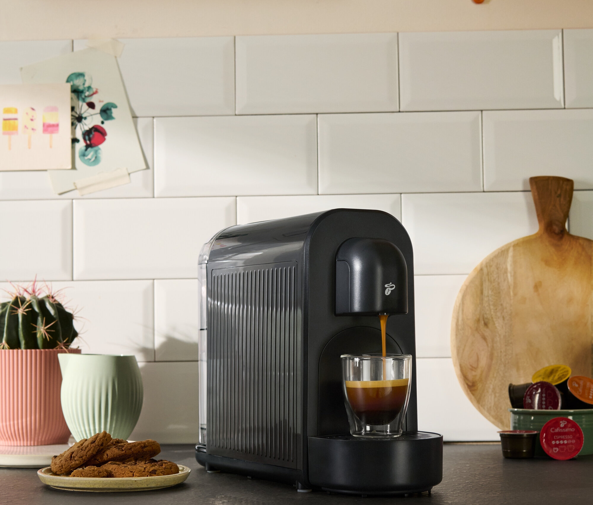 Černý kávovar plní šálek kávou. Vidět jsou také sušenky, sukulent a kuchyňské dekorace.