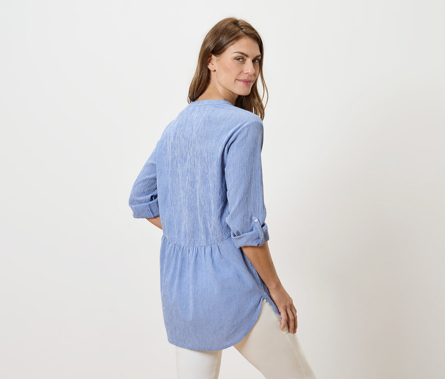 Žena se dívá přes rameno, má na sobě modrou halenku a bílé kalhoty.