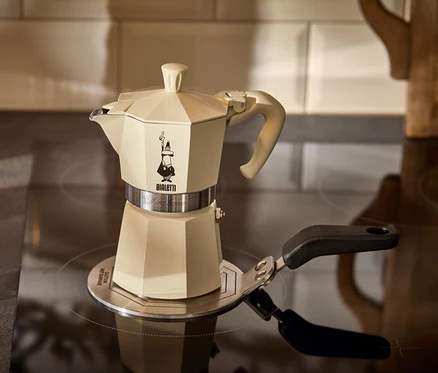 Krémová konvička na espresso Bialetti Moka Express 190 ml stojí na indukční plotýnce Bialetti 13 cm vedle sklenice espressa.