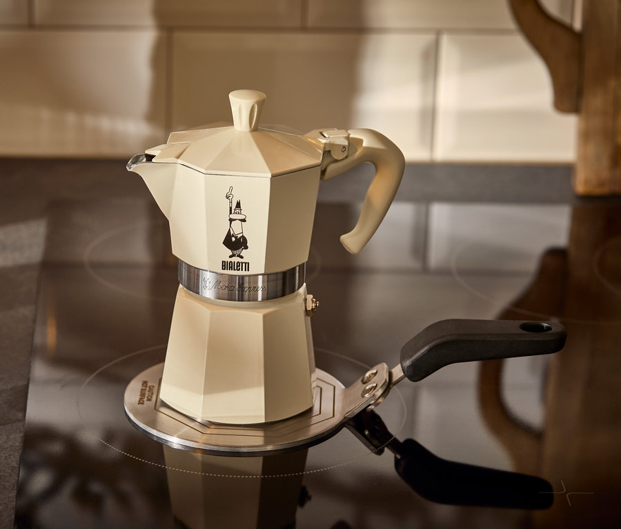 Krémová konvička na espresso Bialetti Moka Express 190 ml stojí na indukční plotýnce Bialetti 13 cm vedle sklenice espressa.