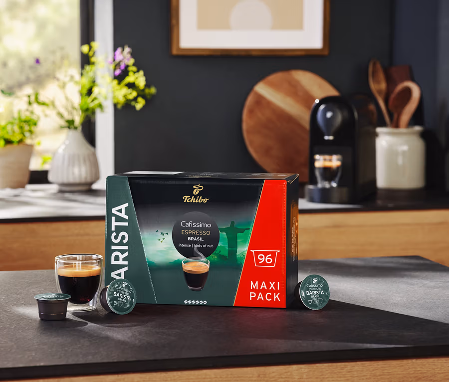 Na stole stojí balení kapslí Cafissimo BARISTA Espresso Brasil, sklenice s espressem a kávovar.