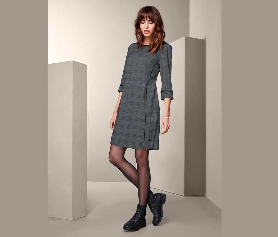 Žena pózuje v šatech z teplákoviny s tříčtvrtečními rukávy a černých botách.