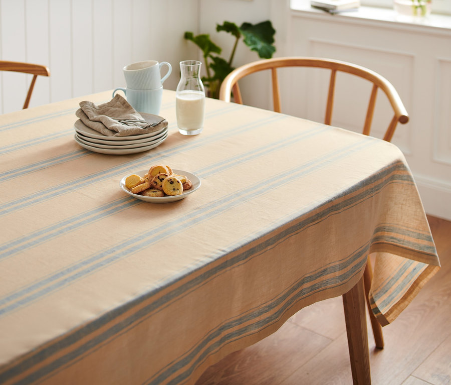 Prostřený stůl s proužkovaným lněným ubrusem standardní velikosti, na němž jsou talíře, šálky, sklenice mléka a sušenky.