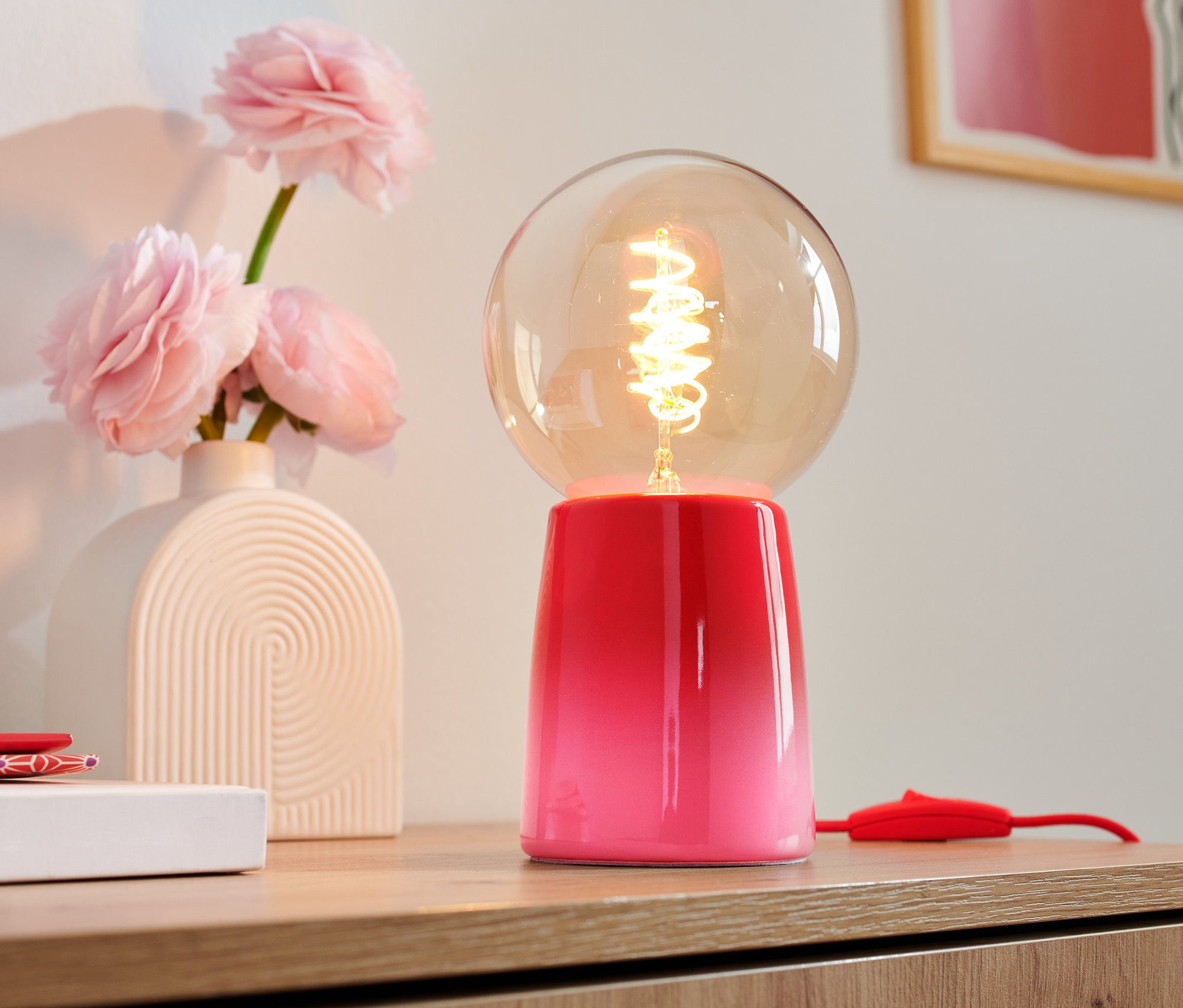 Na dřevěné komodě stojí LED stolní lampa s kulatým skleněným stínidlem a růžovou základnou, vedle vázy s růžovými květinami.