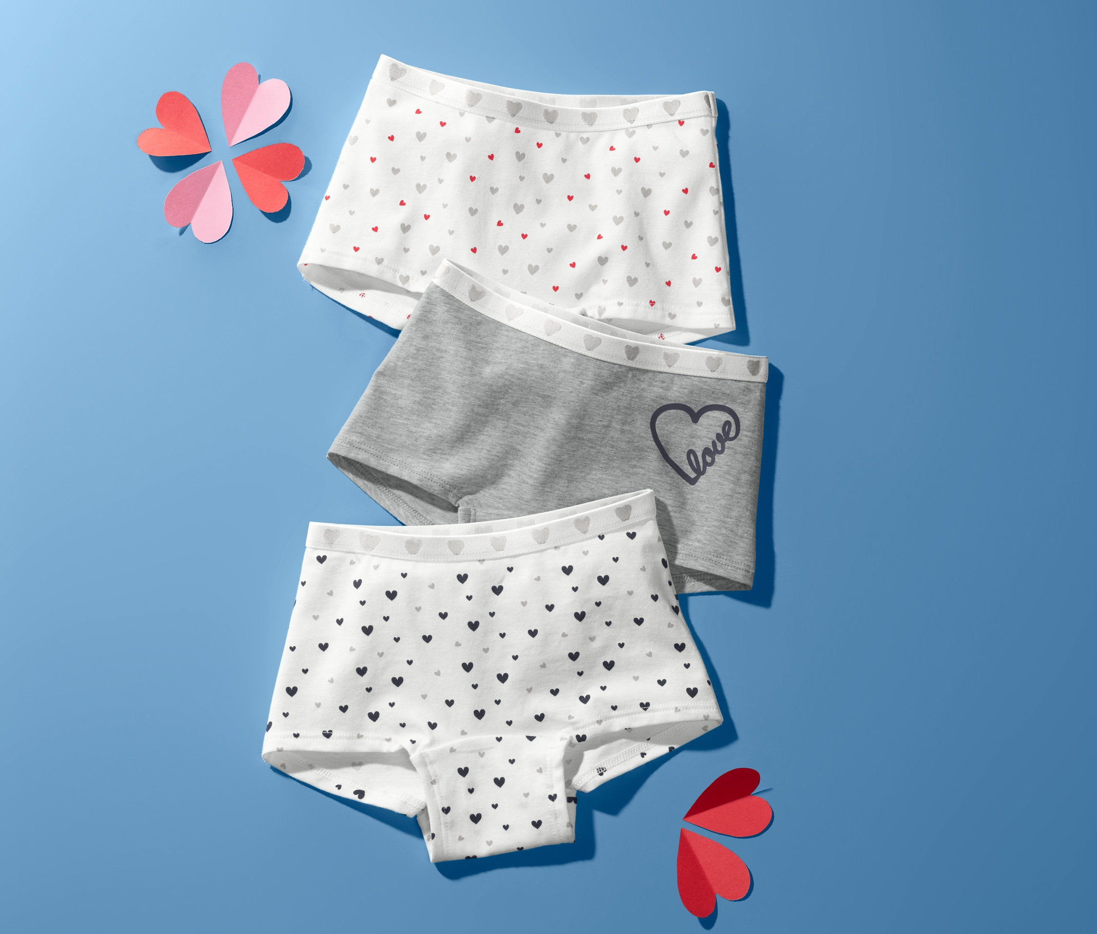 Na modrém pozadí leží troje dívčí kalhotky a ozdoby ve tvaru srdce.