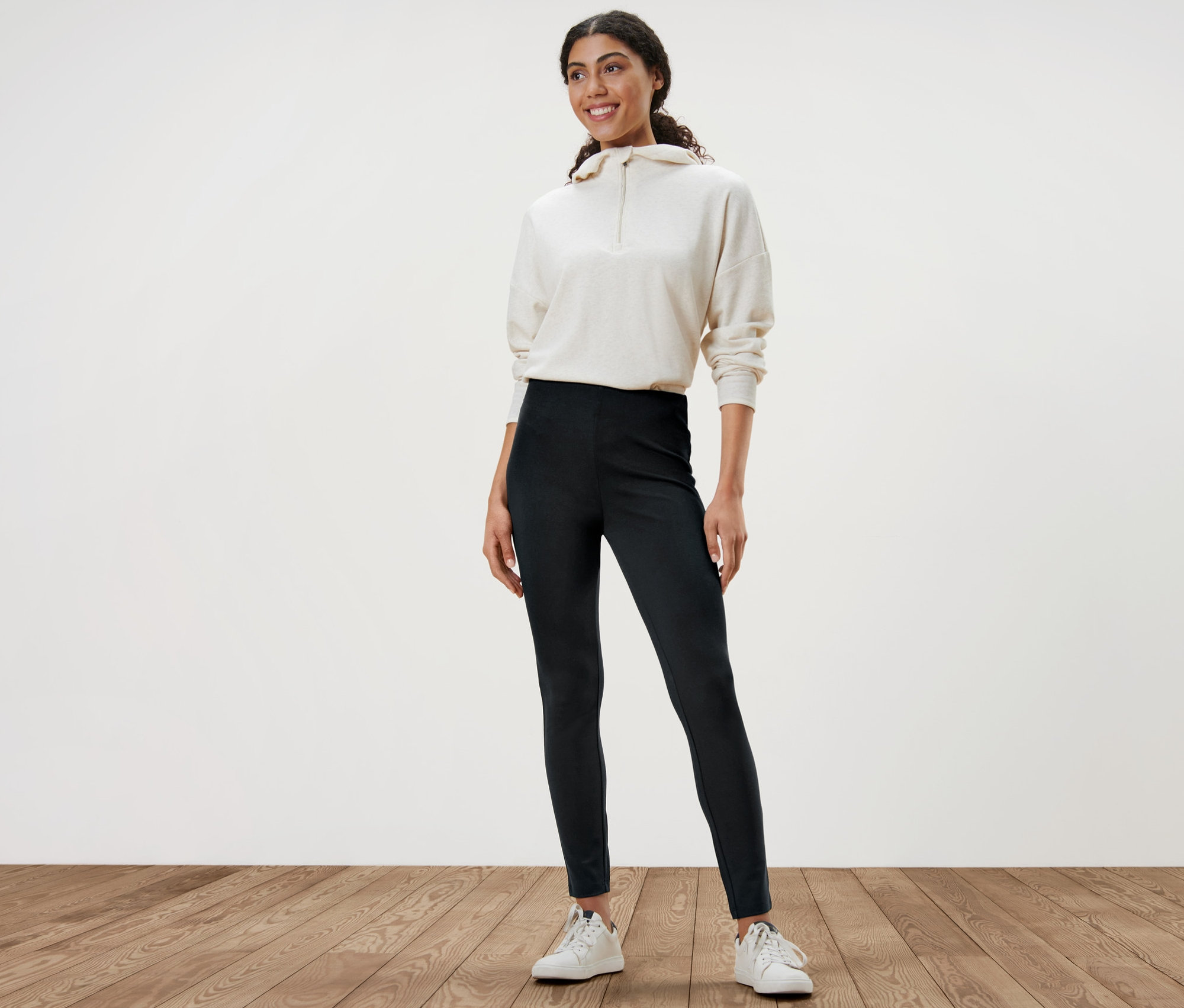 Žena stojí a má na sobě černé pevné legíny, bílou mikinu a bílé tenisky.