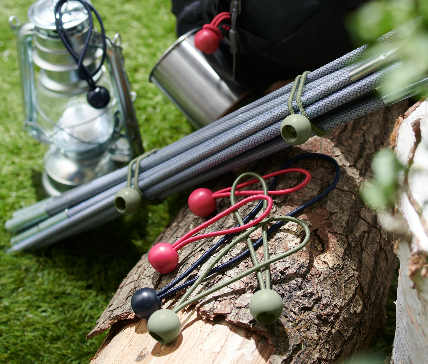 Na kládě leží 12 upínacích gum s kuličkou, vedle nich je lucerna, kovový hrnek, tyče a batoh.