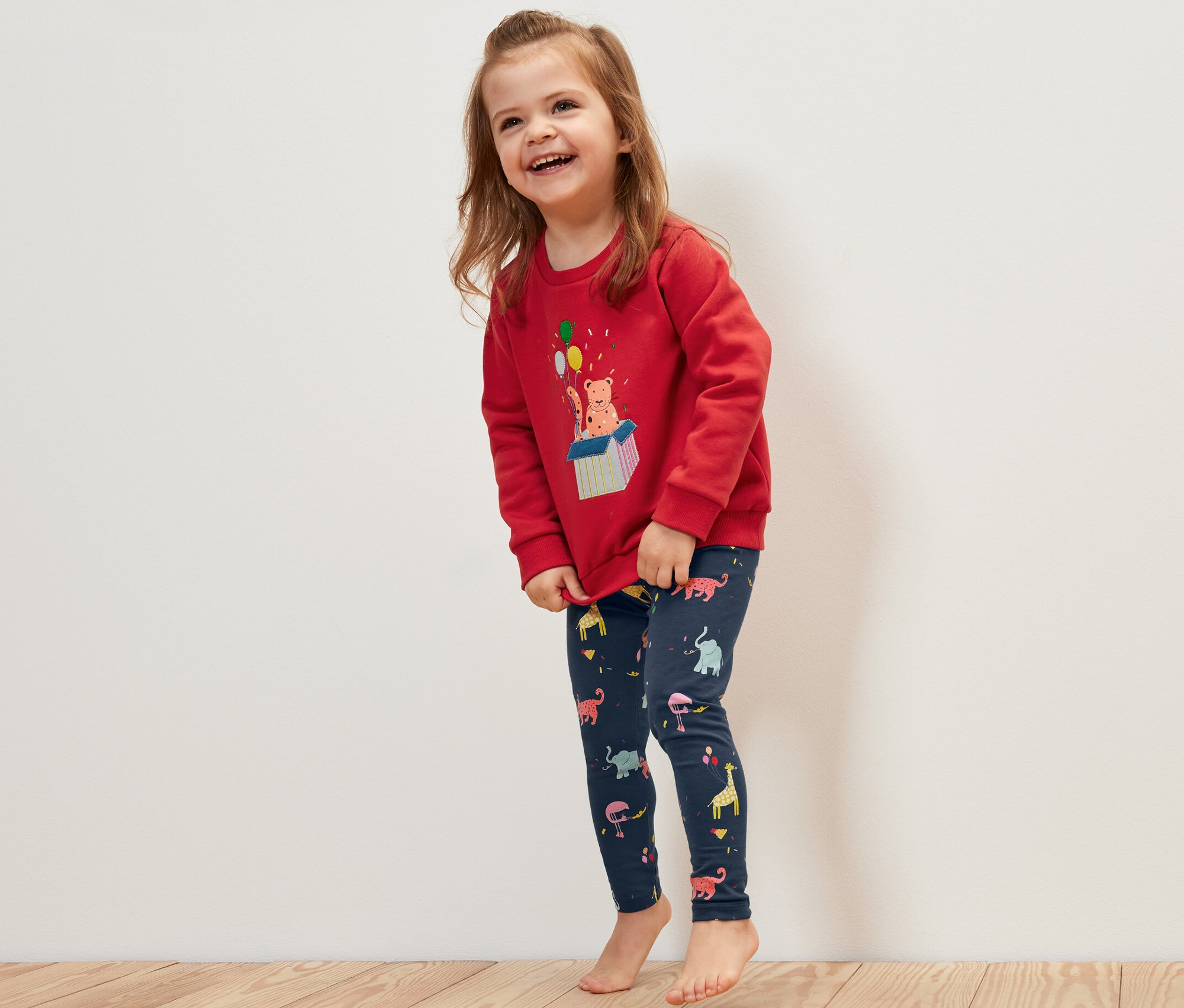 Usmívající se holčička má na sobě červenou mikinu a dětské legíny s potiskem zvířat, 5 ks.