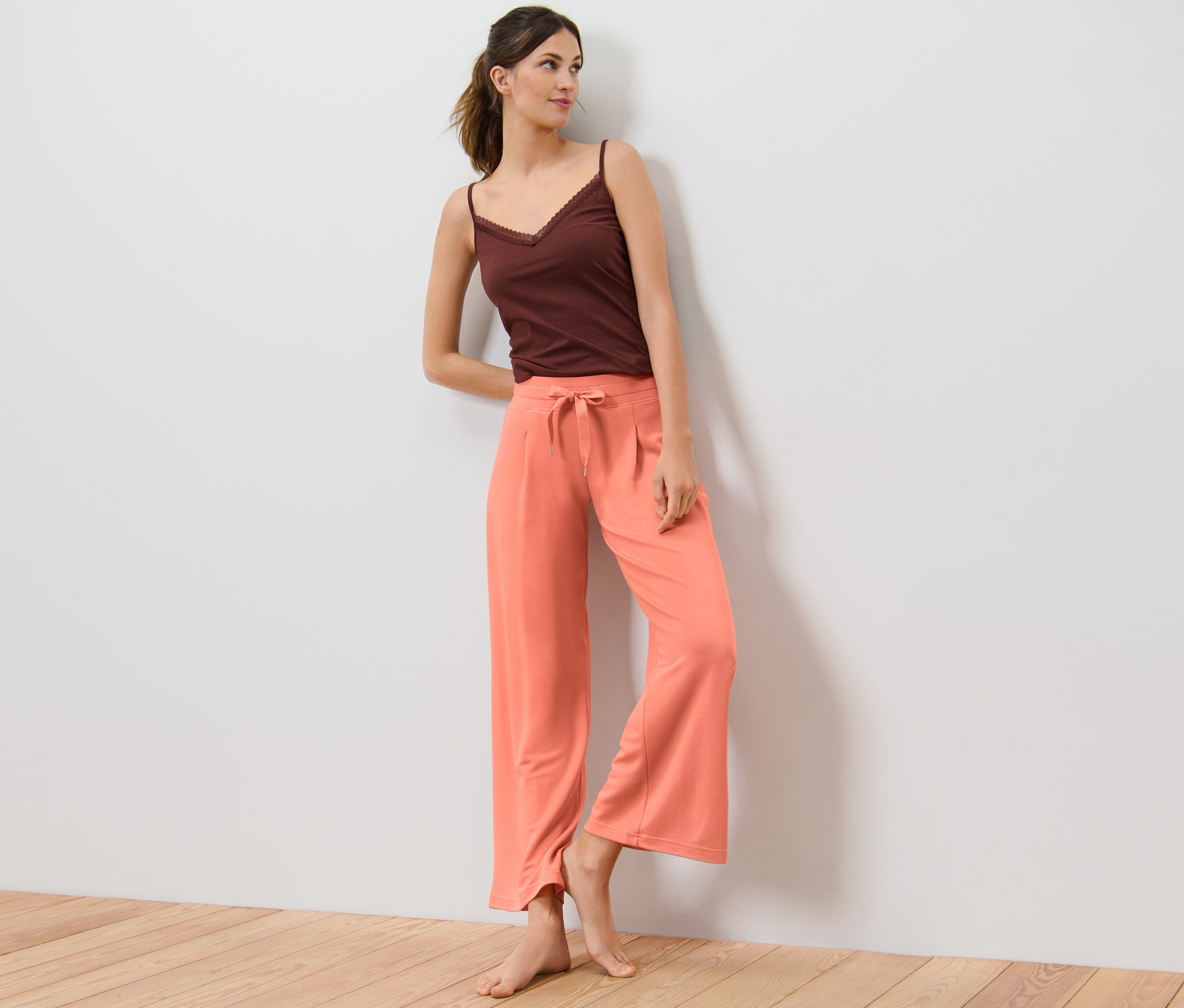 Žena opřená o zeď v pohodlných kalhotách a hnědém topu.