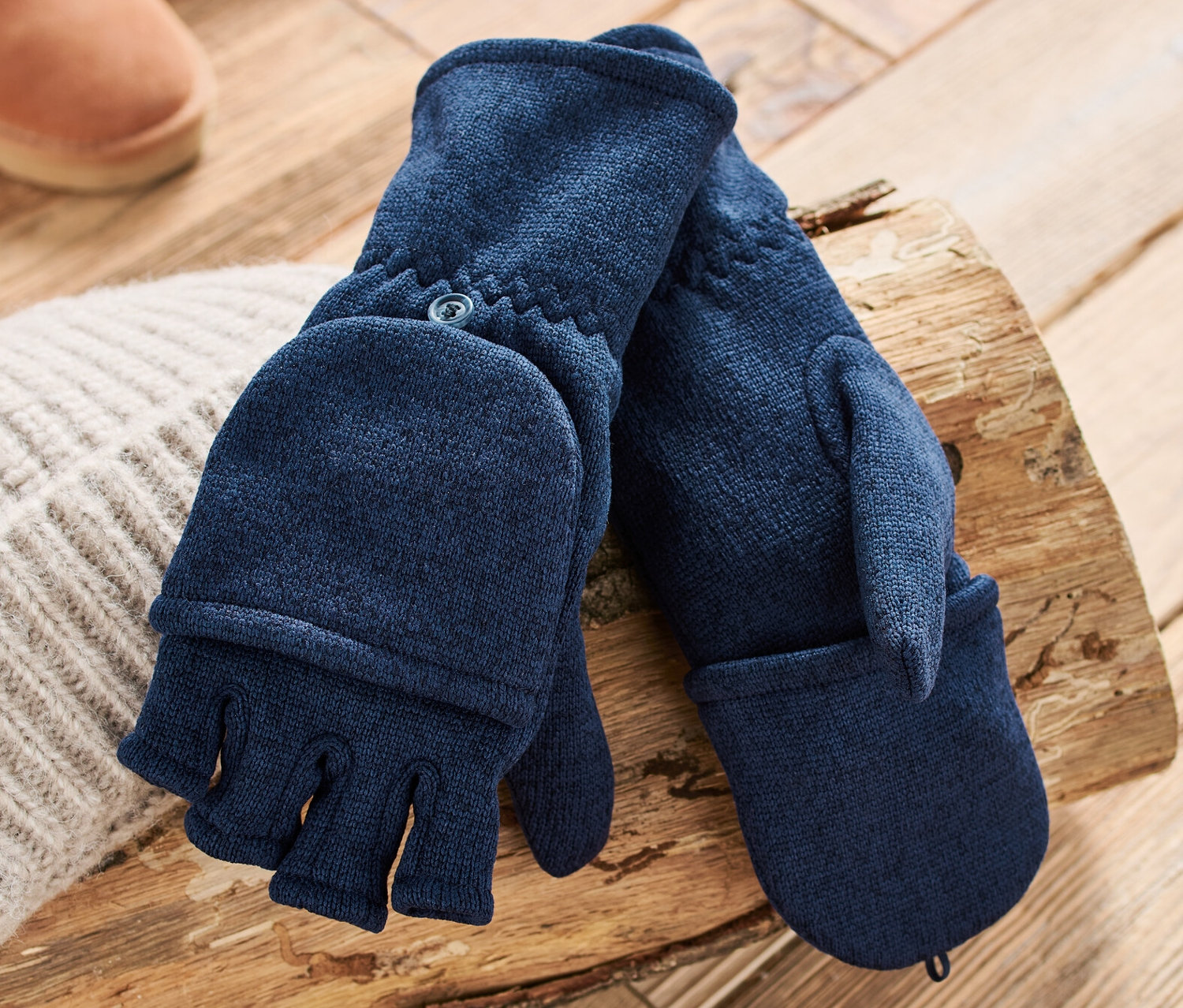 Tmavě modré odklápěcí palčáky z pleteného fleecu leží na dřevěném špalku vedle pletené čepice a hnědých bot.