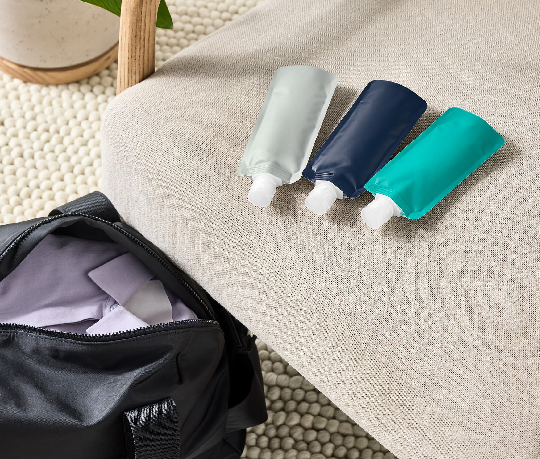 Na béžovém křesle leží tři doplňitelné cestovní láhve vedle černé sportovní tašky a rostliny v květináči.